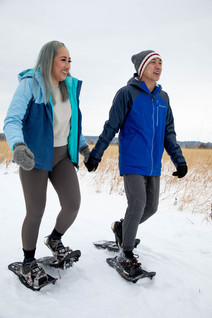 Millennial Asian woman and man holding hands and smiling while snowshoeing on trail through the prairie.