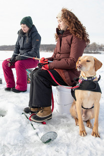 Two Millennial women ice fishing. Brown dog is sitting by them.