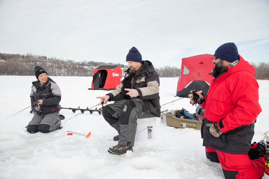 An Asian woman, a white man and a Latino man ice fishing together. Fishing shanty can be seen in the background.
