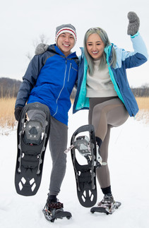 Millennial Asian couple in snowy prairie lifting feet with snowshoes and laughing. 