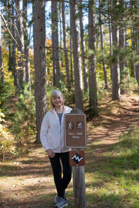 Woman standing by a sign on a hiking trail, with tall trees in the background.