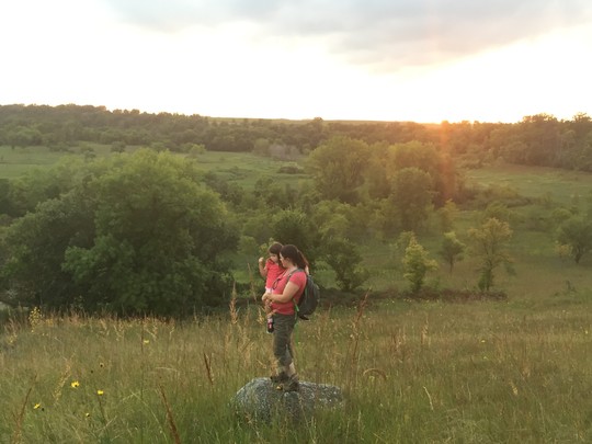 Woman standing on boulder in the middle of a green prairie holding young child in her arms at sunset.