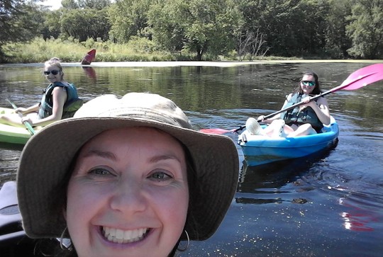 Selfie of woman smiling big with two young women on sit-on-top kayaks posing behind her on the river.