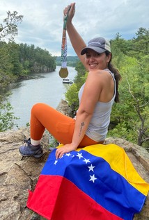 Woman sitting on rock with a Venezuelan flag draped over it. She's holding and showing a medal. River and bluffs are shown in the background.