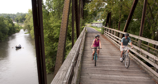 A woman and man biking on a bridge on a trail, a kayaker can be seen on the side in the river.