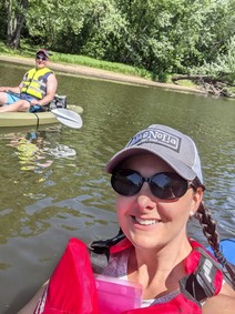 Selfie of woman in life jacket in a kayak in the river, a man on a sit-on-top kayak is posing in the background. 