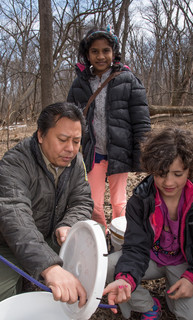 Hmong man demonstrating the first stage of the maple syruping process for two young BIPOC girls.