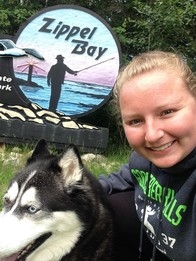 Selfie of blonde woman smiling with husky posing with a sign that reads Zippel Bay and has a silhouette of an angler.