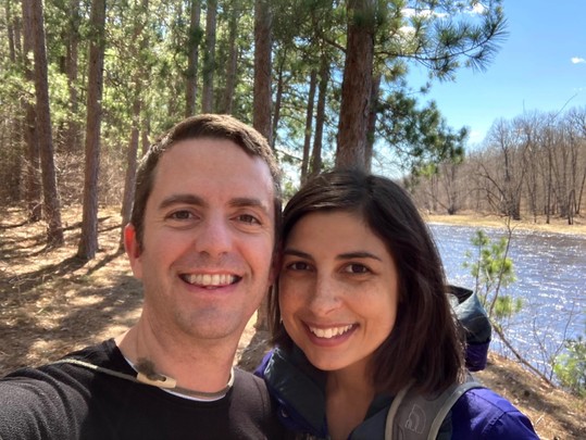South Asian woman with her husband smiling at the camera during a hike through the woods with the river in the background.