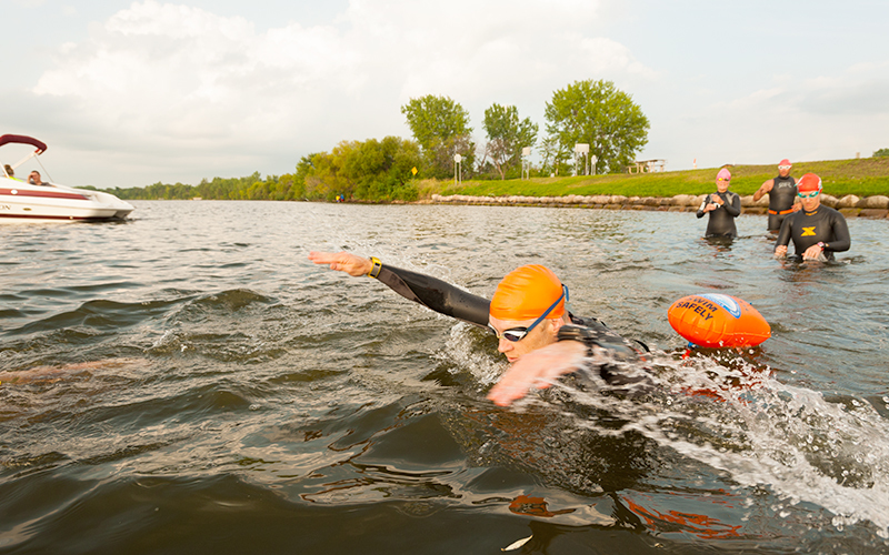 Swimmer with cap and goggle in lake. In the background, a motorboat and other swimmers. 