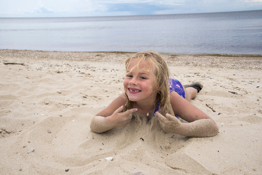 Young blond child laying on the beach and playing with sand, blue skies and big lake seen in the background.