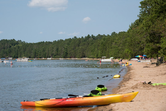 Lake beach surrounded by trees. Kayaks on shore, colorful umbrellas and beach-goers. A couple of pontoons can be seen on the lake.