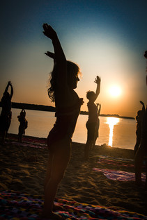 People practicing yoga on the beach at sunset. Orange sun is reflected in the peaceful lake water.