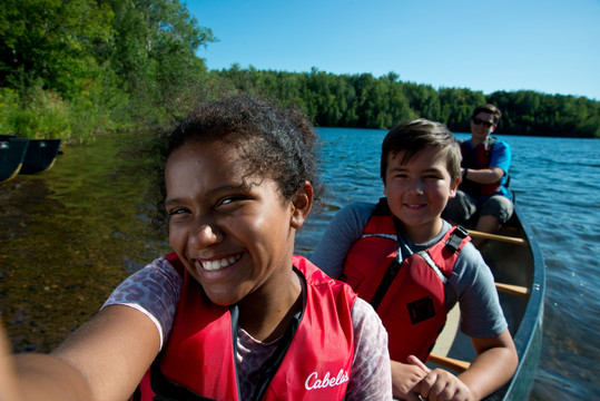 A mixed race youth taking a selfie while floating in a canoe with a white boy and a young white adult.