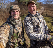 two archery hunters in camo gear holding bows near a road