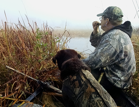 duck hunter blowing on a call with a dog next to him in a marsh