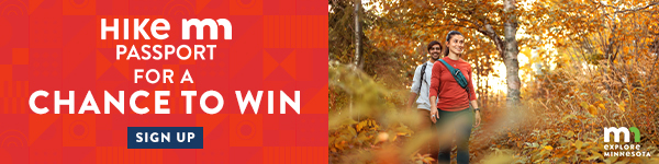 Text reads, "Hike MN Passport for a chance to win. Sign up. Explore Minnesota." Photo shows two people walking through the woods in fall.  