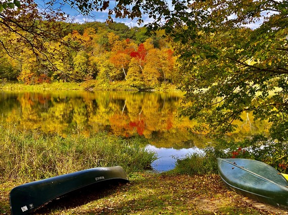 Two canoes upside-down by a lake. Fall colors are everywhere on trees, grasses, and the reflection on the quiet water.