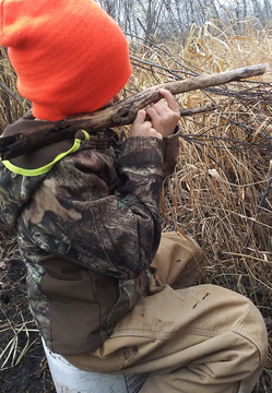 kid in a blaze orange hat sitting on a bucket in woods