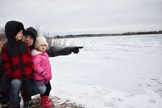 A dad and his two kids; the dad is pointing at a frozen lake