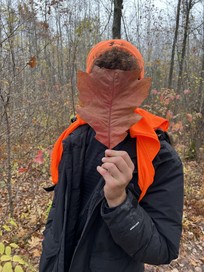 Young person holding a large leaf in front of their face. You can see the blaze orange beanie and accents on the jacket for safety.