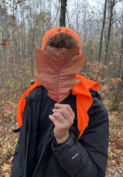 Young person holding a large leaf in front of their face. You can see the blaze orange beanie and accents on the jacket for safety.