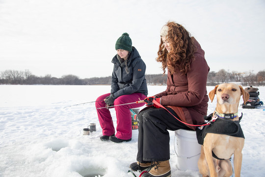 Two young women ice fishing. Their puppy is sitting by them.