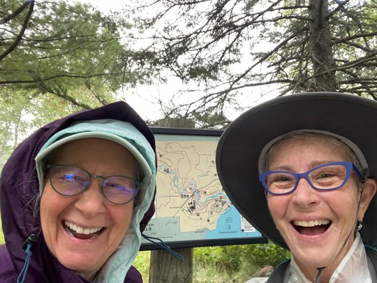 A selfie of two very happy, smiling female hikers posting with map sign for Temperance River State Park.
