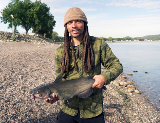 Young Black man with dreads standing on a lake beach on a sunny day, holding a large fish.