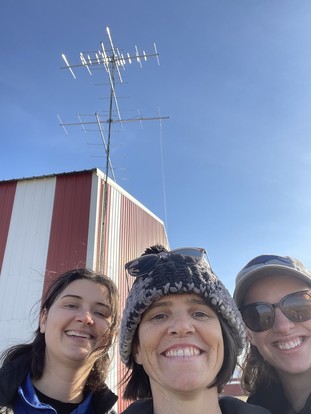 three white women smiling with a large metal antenna behind them