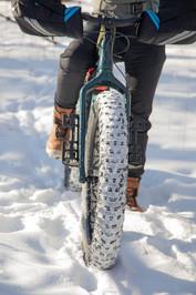 Close-up of a bike's fat tire covered in snow. Rider's legs can be seen in the background. 