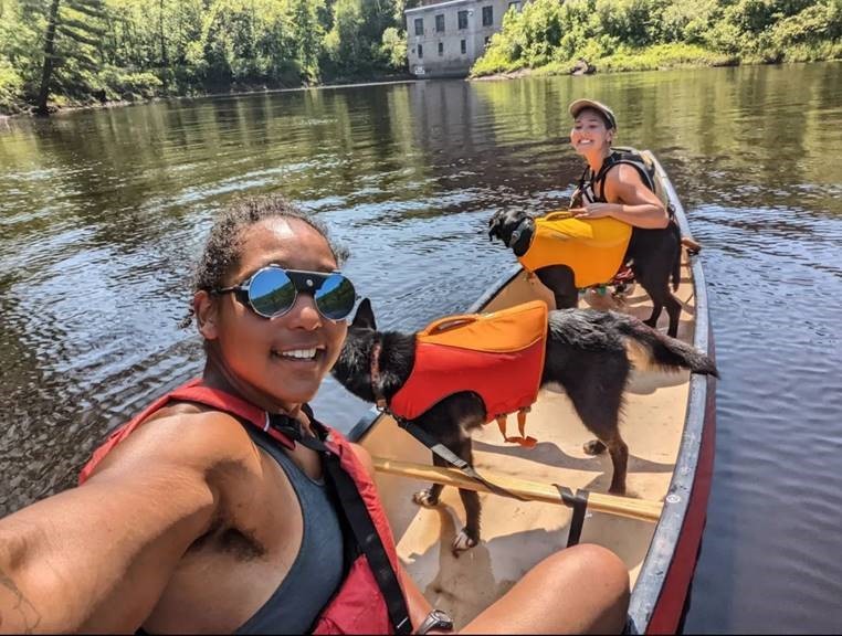 A Black woman, a Japanese American woman and two dogs enjoying a canoe outing on a sunny day.