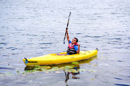 Young Black child floating in a yellow kayak holding the paddle up in the air.  