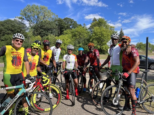 A group of cyclists posing with their bikes, some sporting Major Taylor Cycling Club bike jerseys. Green trees and blue skies in the background.