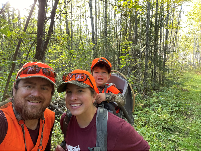 Two people in the woods, with the woman wearing her son in the harness on her back