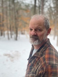 Man with a beard and plaid shirt looking at the camera, a snowy forest can be seen in the background.