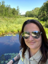A woman in park ranger uniform, wearing sunglasses and posing by a lake. 