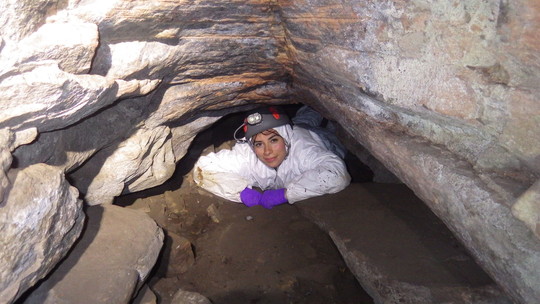 Melissa Boman in protective gear, laying on the ground in a small tunnel in a cave