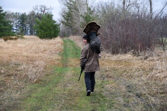A woman looking back over her shoulder at the camera but walking forward on a trail, holding a gun and a turkey on her other shoulder