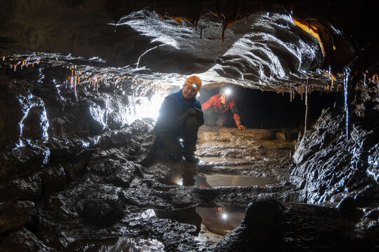 Two cavers in full gear traveling through a muddy cave passage.
