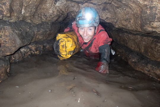 Caver in full gear going through a narrow, muddy passage while holding a yellow dry bag.