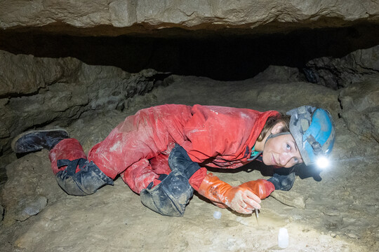 Female researcher laying on cave floor collecting water from a puddle.