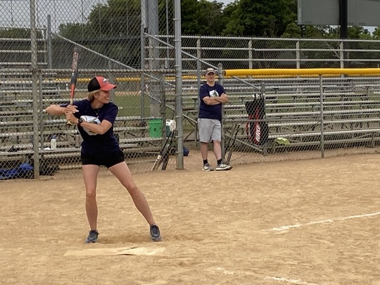 A woman prepares to swing a bat on a softball field, with an onlooker in the background