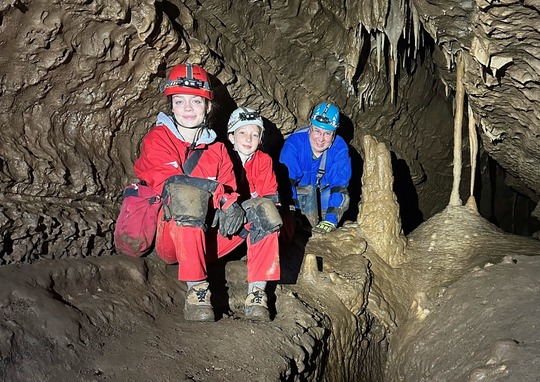 Three people in a cave, wearing red and blue suits and headlamps 