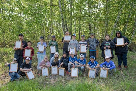 A class of children hold handmade lap desks in a forest