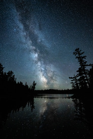 Milky Way reflected on a quiet lake, framed by trees.