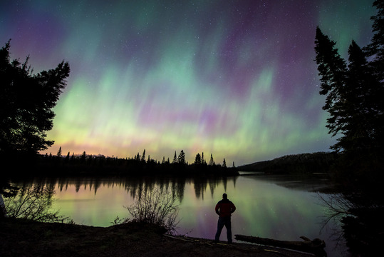 Photographer standing by a lake while the northern lights are glowing purple and green, reflecting in the water.