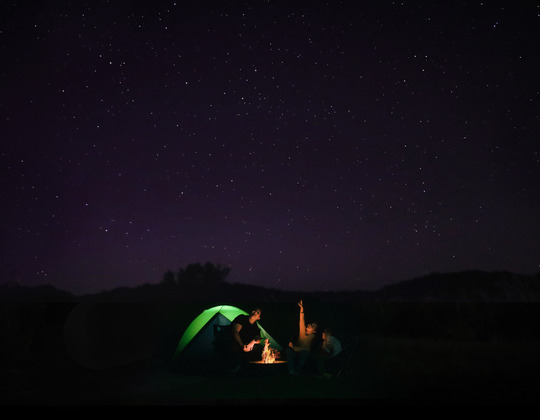 Mom and two young daughters at campfire, looking and pointing at the stars. Their dimly lit tent is seen in the background.