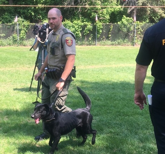 A man in a conservation officer uniform holds the leash of a black dog