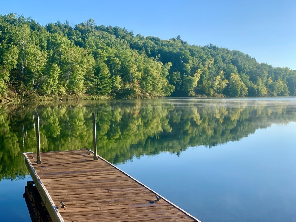 A quiet lake reflecting trees on shore and a blue sky. A dock is seen in the foreground, providing depth to the photo.